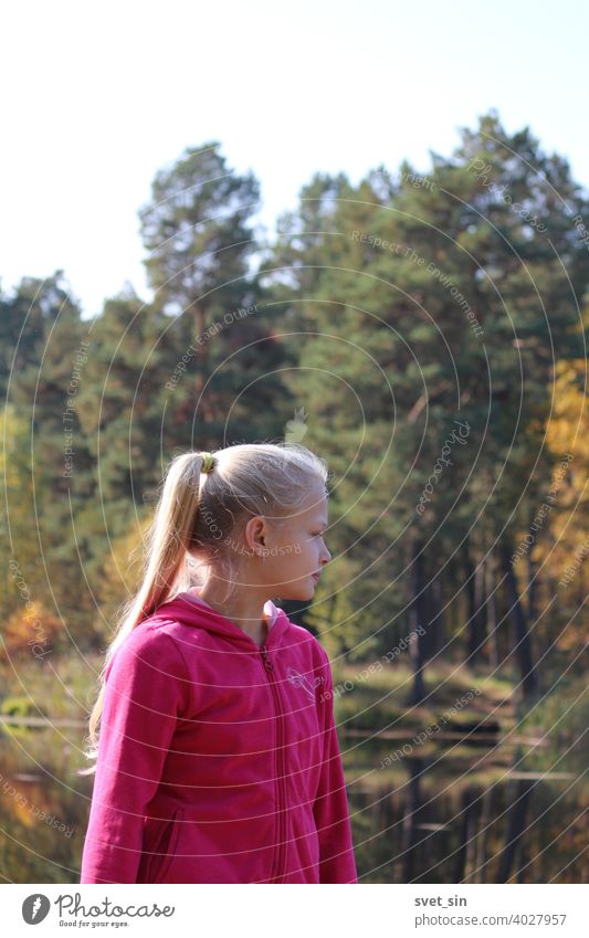A teenage girl with long blond hair stands on the shore of a lake against the backdrop of water and autumn yellow forest in autumn sunlight. Portrait of a blond girl in profile.