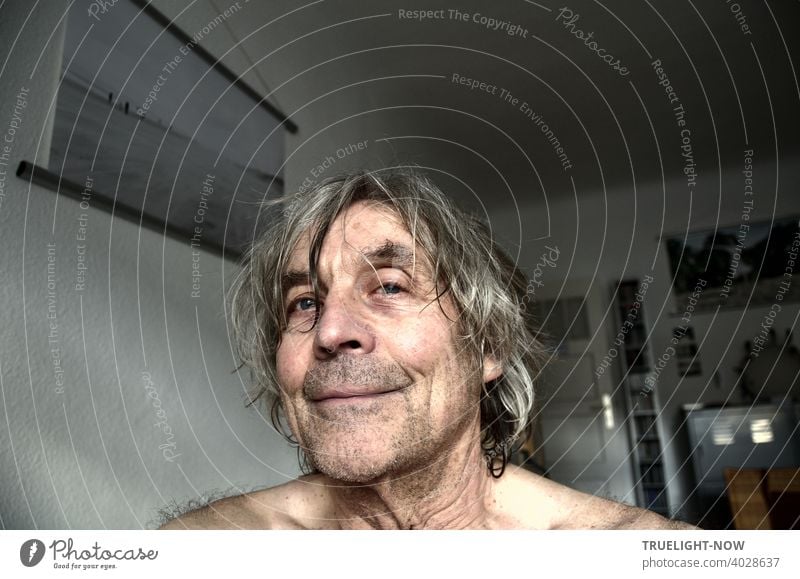 Frontal portrait of a senior citizen in his home office, who seems pleased that he hasn't been able to visit the hairdresser in over a year because of the pandemic. On the wall a photo of the Photocase user CL