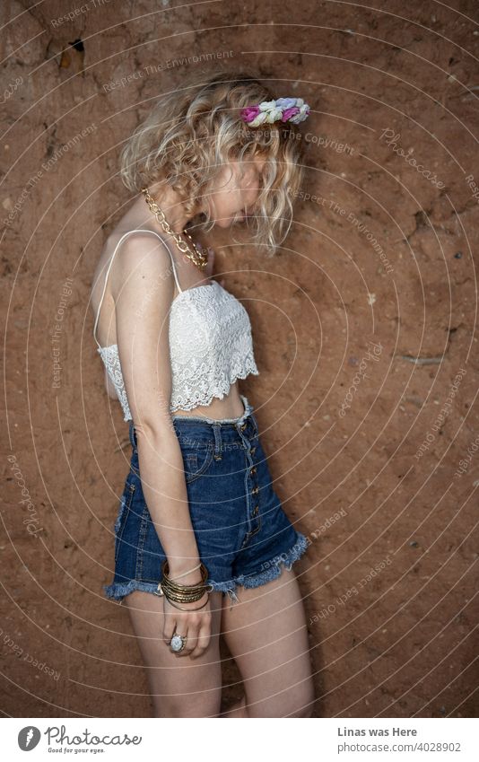 Model test of a gorgeous blonde girl dressed in a white blouse and blue shorts. Curly blond hair is hiding her face. A mysterious portrait against the wall.