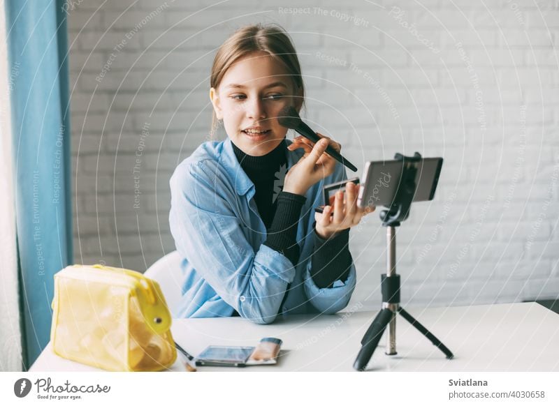 A teenage girl does makeup and records video for her blog on a smartphone on a tripod at home. Communication of a video blogger girl on social networks with subscribers and friends