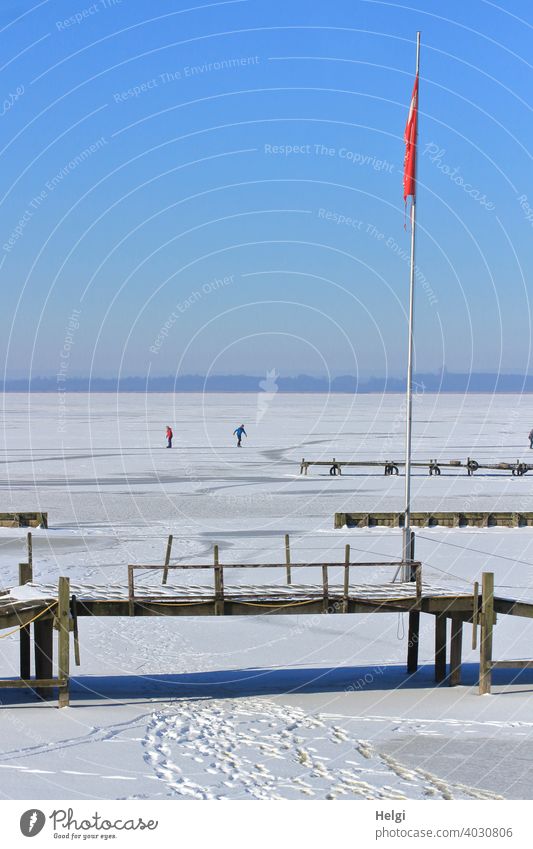 frozen lake with snow, tracks, wooden walkways, a flagpole and in the background two people skating Winter Snow Frost Ice chill Footbridge Tracks skate