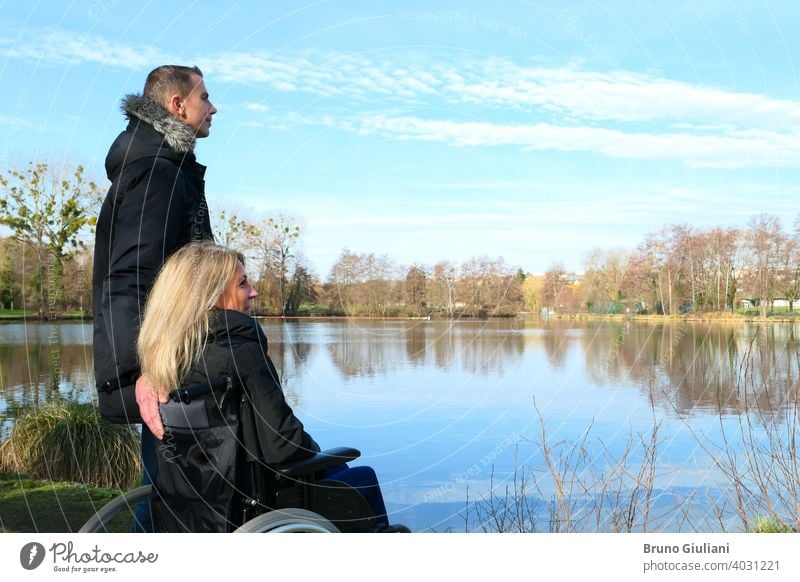 Concept of disabled person. A woman in a wheelchair with a man standing next to her, outside in the nature in front of a lake adult concept contemplation couple