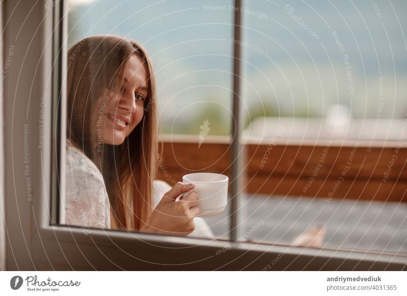 Young female standing after taking a shower in the morning on balcony of the hotel. holding a cup of coffee or tea in her hands. Looking outside nature forest and Mountain