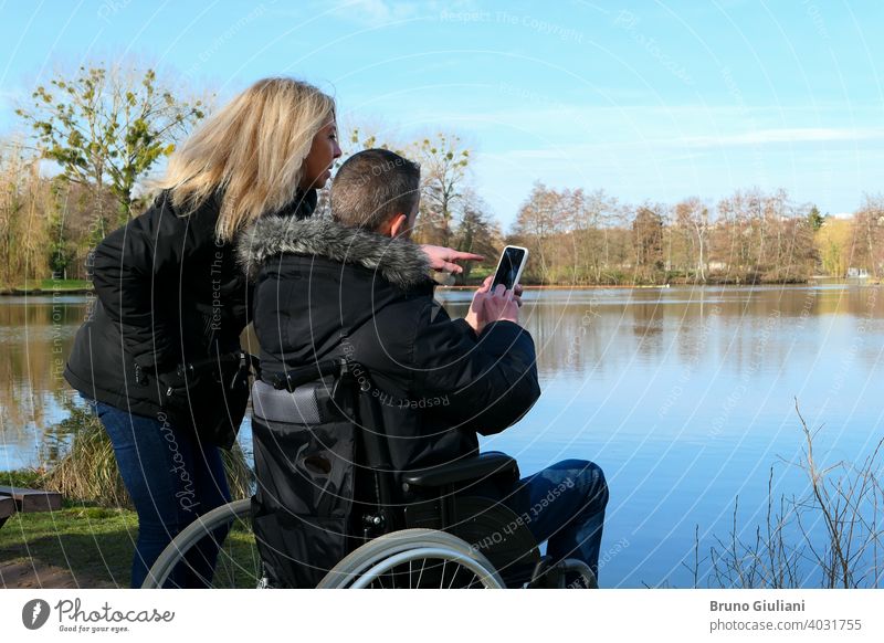 Concept of a person with a physical disability. A man in a wheelchair with a woman standing beside him. Couple using technology while looking at a smartphone. Rural scene by a lake.