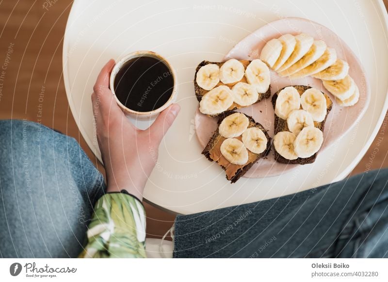 Woman holds a cup of black coffee, shot directly above. Having healthy fruit and peanut butter sandwich for breakfast, drinking morning coffee at home banana
