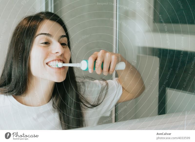 A young woman with black hair brushing his teeth with an electrical toothbrush hygiene dental bathroom female person smiling happy indoor healthcare oral smile