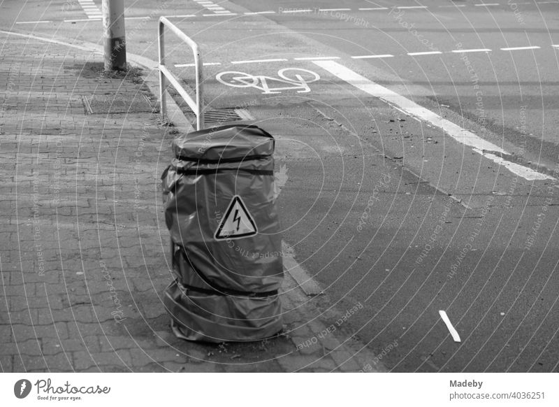 Warning of high voltage on a pillar wrapped with foil between sidewalk and bicycle path in the Westend of Frankfurt am Main in Hesse, photographed in neo-realistic black and white
