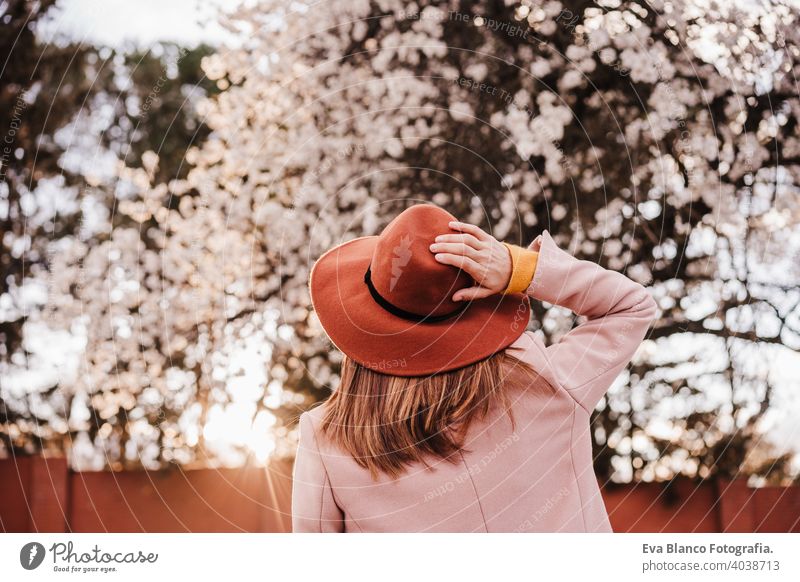 back view portrait of young woman wearing modern hat at sunset in park. beautiful almond tree flowers background. Springtime and blossom spring trees outdoors