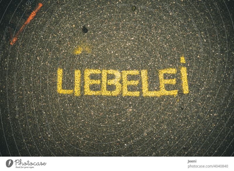 A path with street painting or graffiti with the word "LIEBELEI" | small orange colored stripe on the top left | text on a plain neutral background Love Dating