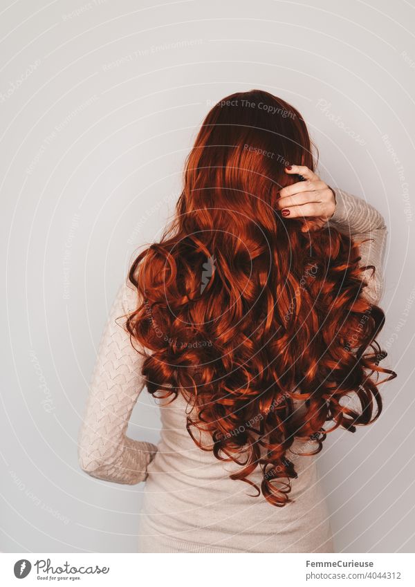 Back view of a woman with long red curly hair in a skin-colored sweater who is grabbing her hair with one hand. Neutral background Copy Space top
