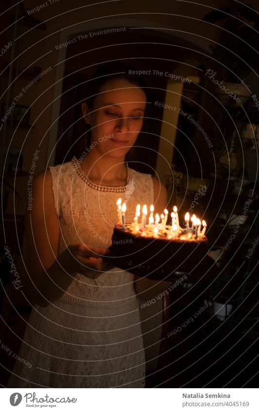 Young girl in white lace dress holding cake with burning candles in her hands in dark room. Birthday celebration at home. Vertical orientation. young woman
