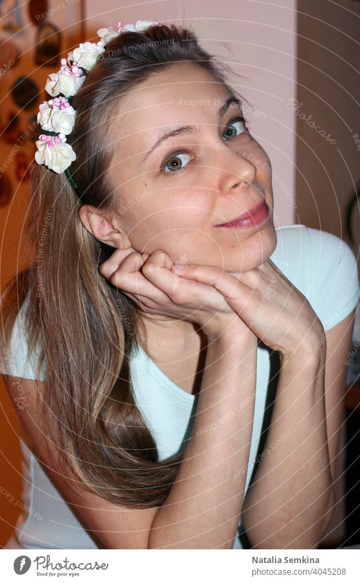 Attractive young girl in light dress and wreath with flowers on her head siting with her head propped on her hands and looking at camera close-up. Vertical orientation.