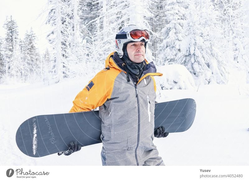 Beautiful caucasian man in a bright yellow and grey jumpsuit, white helmet and goggles is holding his snowboard. Winter sports, leisure activity. Vacation in the mountains. Front view.