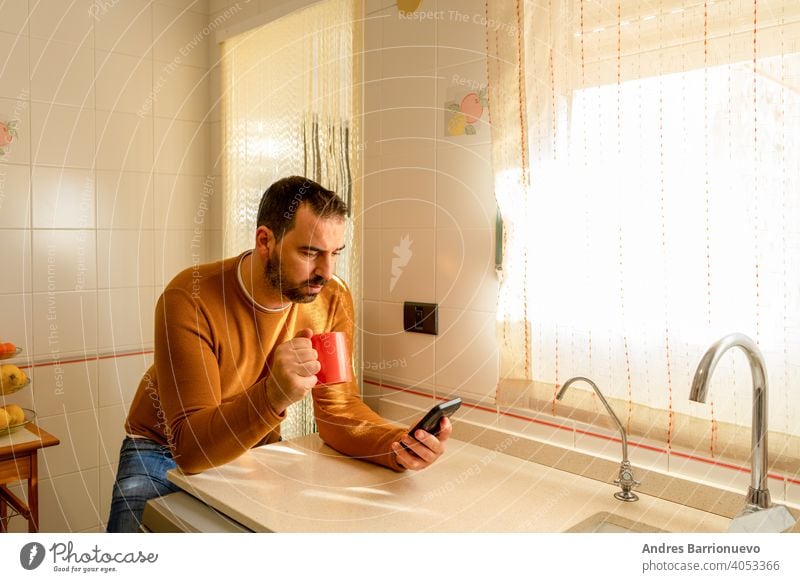 Handsome bearded man in an orange sweater looking at the smartphone in the kitchen while having a cup of coffee mobile phone communication guy drink positive