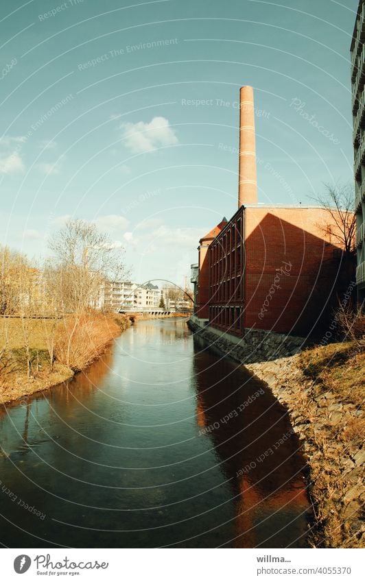 A sunny day by a quiet river with a factory with a smokestack on its banks. Time thinks the little clouds should also be mentioned. River Factory Chimney