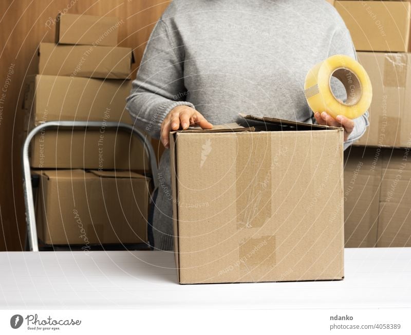 woman in a gray sweater holds a roll of duct tape and packs brown cardboard boxes on a white table, behind a stack of boxes lifestyle real adhesive tape many