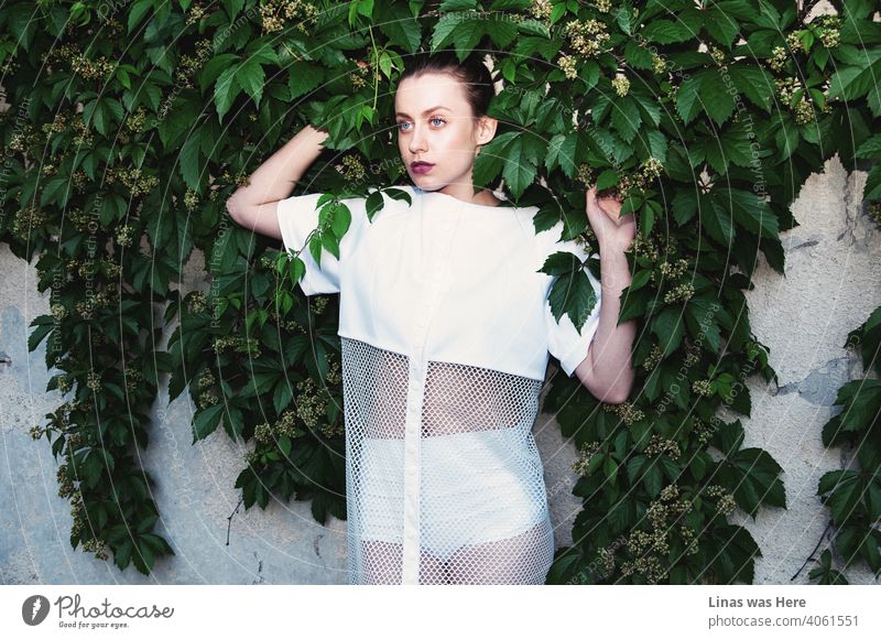 Brunette fashion model is posing with her avant-garde dress next to green creepers. Her white outfit is looking fashionable as hell. The future is now!