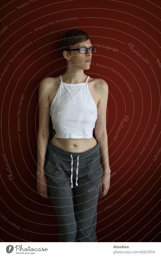 Young woman in with belly top and sweatpants stands in front of red wall and looks out of window Woman Slim jeans Top Eyeglasses sits Shoulders Skin Large