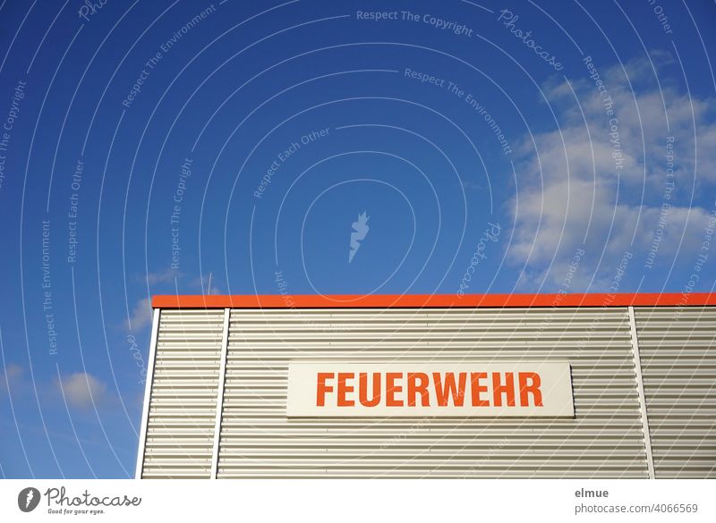 Partial view of a functional building with metal cladding and the inscription in red "FEUERWEHR" in front of a blue sky with fair weather clouds / rescue service / emergency call 112