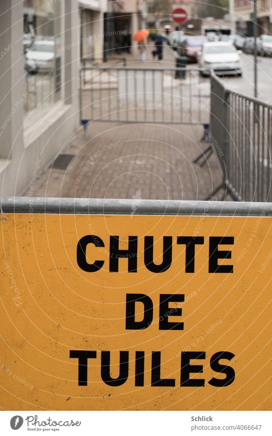 In front of a barrier, a yellow sign in French reads CHUTE DE TUILES Roofing work or storm damage Chute de Tuiles Storm damage cordon Yellow off Exterior shot