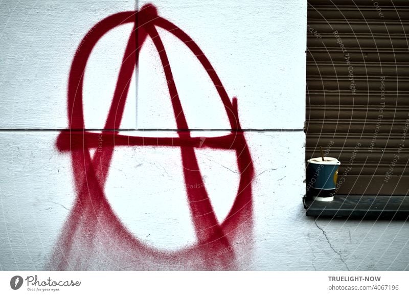 A sprayer has sprayed the well-known signet of the anarchists or anarchos on a white house wall, large and in red color: large A in a circle. The coffee mug on the windowsill next to it in front of a closed brown shutter seems appropriate