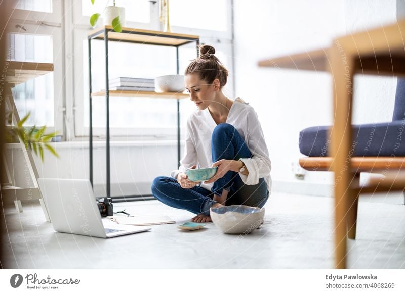 Young female pottery artist using laptop in her workshop internet online computer technology retailer items selling sitting floor shopping millennials student