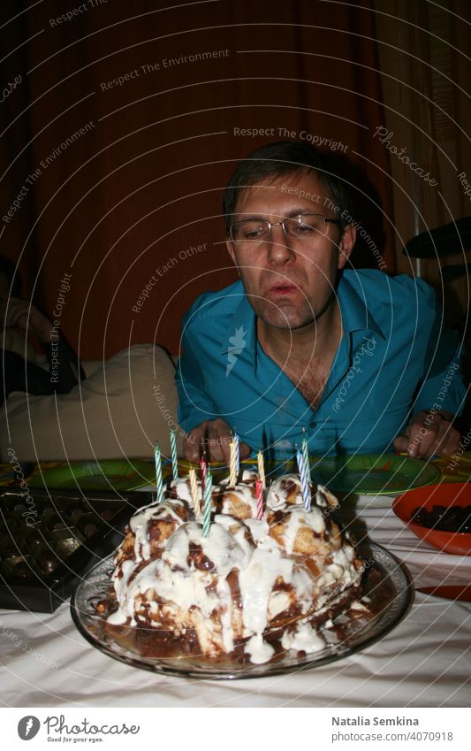 Man in blue shirt blowing out burning candles on  handmade festive cake in dark room. Birthday celebration at home. Party at home. Waist portrait. Vertical orientation.