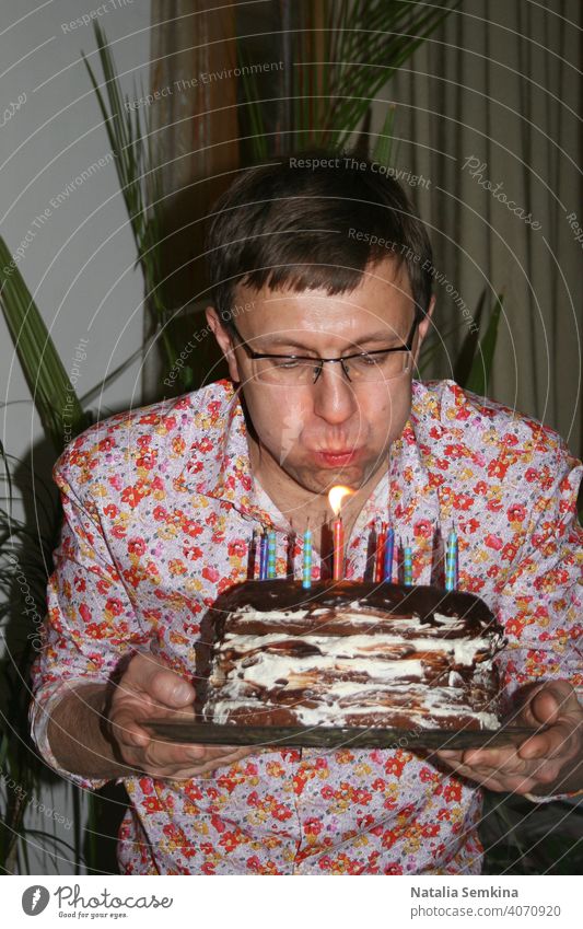 Man in floral shirt holding handmade festive cake in her hands and blowing out burning candles in dark room. Birthday celebration at home. Party at home. Waist portrait. Vertical orientation.