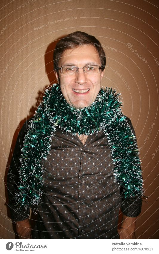 Cheerful smiling man wearing glasses and brown shirt with green tinsel wrapped around his neck standing against a beige wall and looking at camera . Waist portrait. Christmas party at home.