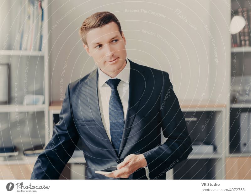 Horizontal shot of handsome male manager in stylish clothes, uses smart phone for checking information about entrepreneurship, reads news in web page, connected to wireless internet, stands at office