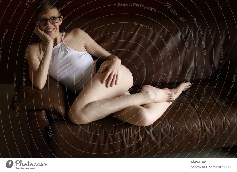 Portrait of a young laughing woman on a brown leather couch Woman Young woman Slim Top Eyeglasses sits Skin Large Athletic Articulated inside Room studio Bright