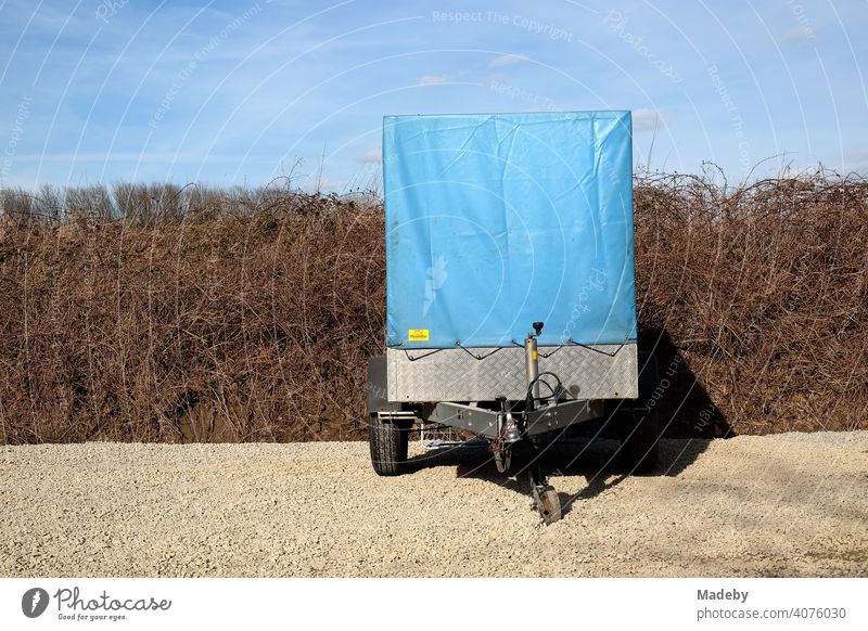 Car trailer with high blue tarpaulin made of plastic in front of brittle hedge and blue sky on gravel in natural colors in Lemgo near Detmold in East Westphalia-Lippe