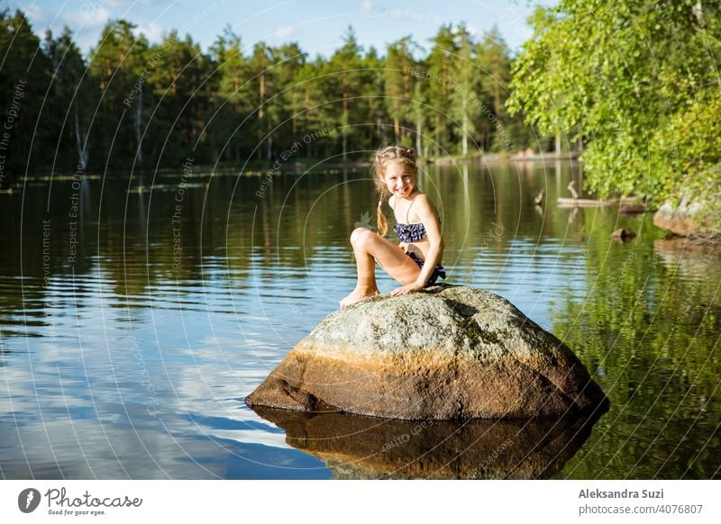 Cute little girl sitting on a rock in lake. Enjoying summer vacation. Child and Nature. Happy isolation concept. Exploring Finland. Scandinavian landscape.