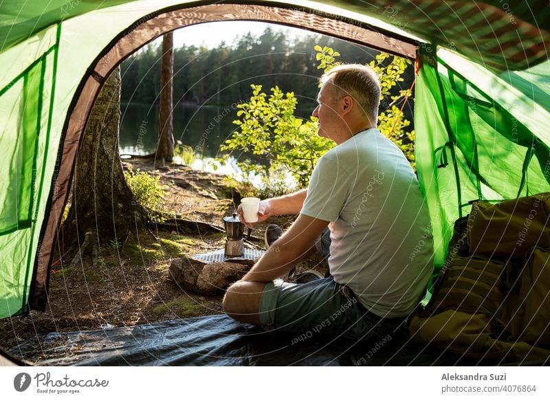 Man making coffee using espresso maker on campfire in forest on shore of a lake, sitting in tent, making a fire, grilling. Happy isolation concept. Exploring Finland. Scandinavian landscape.