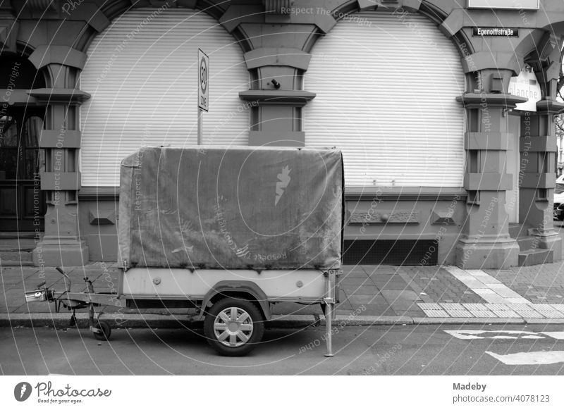 Car trailer with tarpaulin parked in front of a closed restaurant with lowered roller shutter in an old building in the north end of Frankfurt am Main in Hesse, photographed in classic black and white