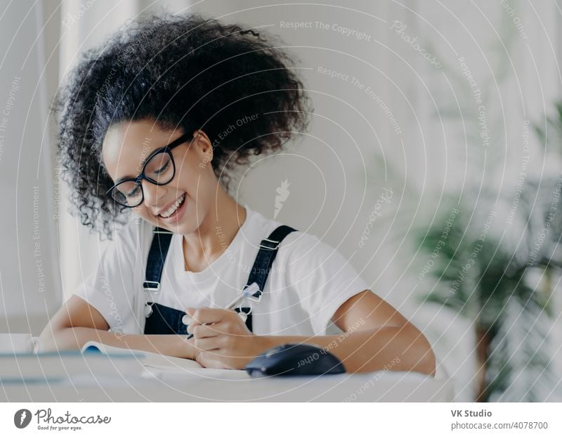 Happy Afro American female student writes down information, prepares for exams, sits in coworking space, has curly dark hair, wears optical glasses white t shirt and overalls, studies in spacious room