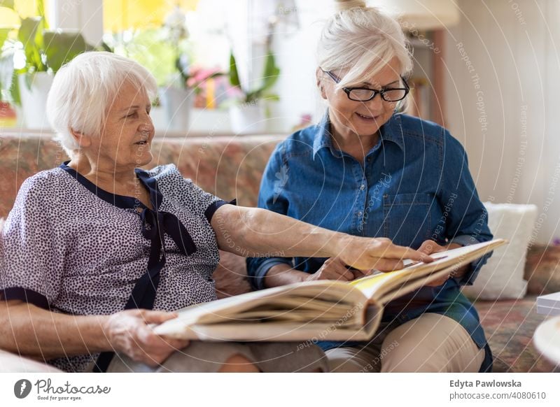 Senior woman and her adult daughter looking at photo album together on couch in living room sitting memory memories nostalgia history photos showing remember