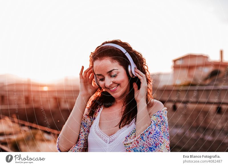 portrait outdoors of a young beautiful woman at sunset listening music on headset and smiling. Lifestyle and music concept casual fun young adult app