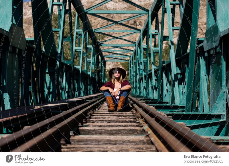 portrait of a young beautiful woman sitting on the railway of a green bridge. Wearing stylish clothes and a hat. LIfestyle. Outdoors. Sunny train female pretty