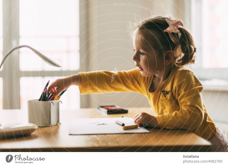 A little girl draws at the table with colored pencils at home or in kindergarten. Childhood, creativity, education. Side view paper cute desk child kid