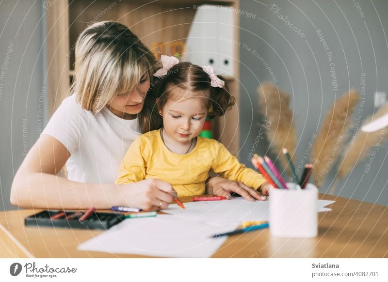 Happy mother and daughter draw together with colored pencils at the table in the room. Time together, creativity, education girl child mom drawing paint paper