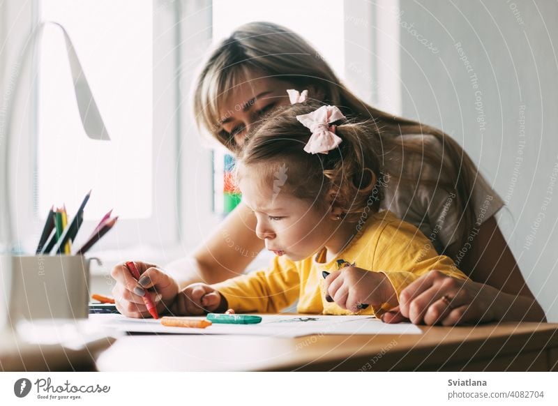 A young mother teaches her little daughter to draw with colored pencils. Time together, creativity, education. Side view girl child mom drawing paint table