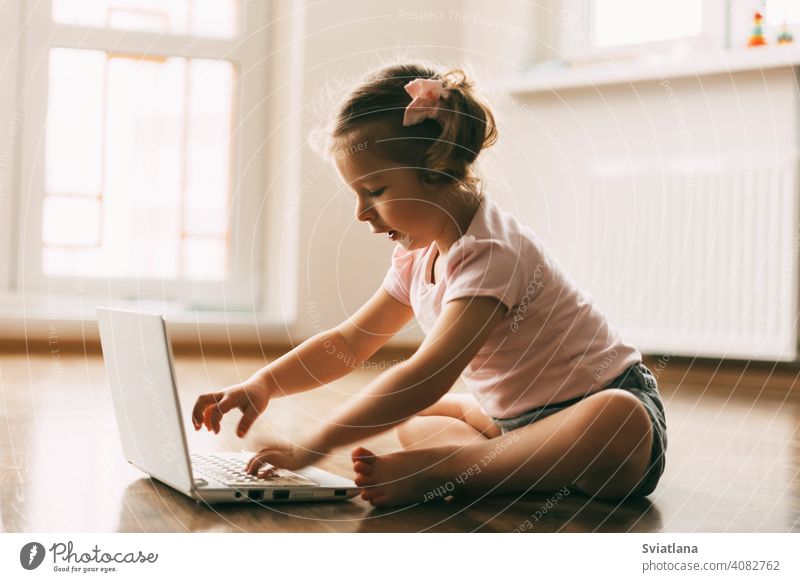 A little girl is playing or watching cartoons on a laptop and sitting on the floor in a room. Childhood, recreation, cartoon time child computer looking online