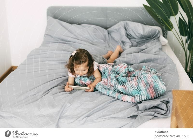 A little girl watches cartoons on her phone or plays on the bed before going to bed. Top view smartphone child kid mobile home small communication looking