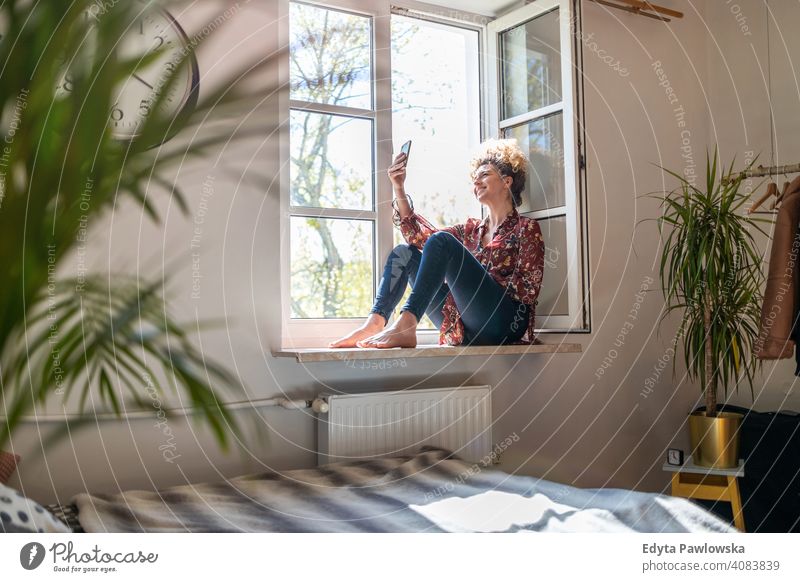 Young woman sitting on window sill and using smartphone happy smiling cheerful apartment leisure bedroom house home alone people caucasian adult indoors person