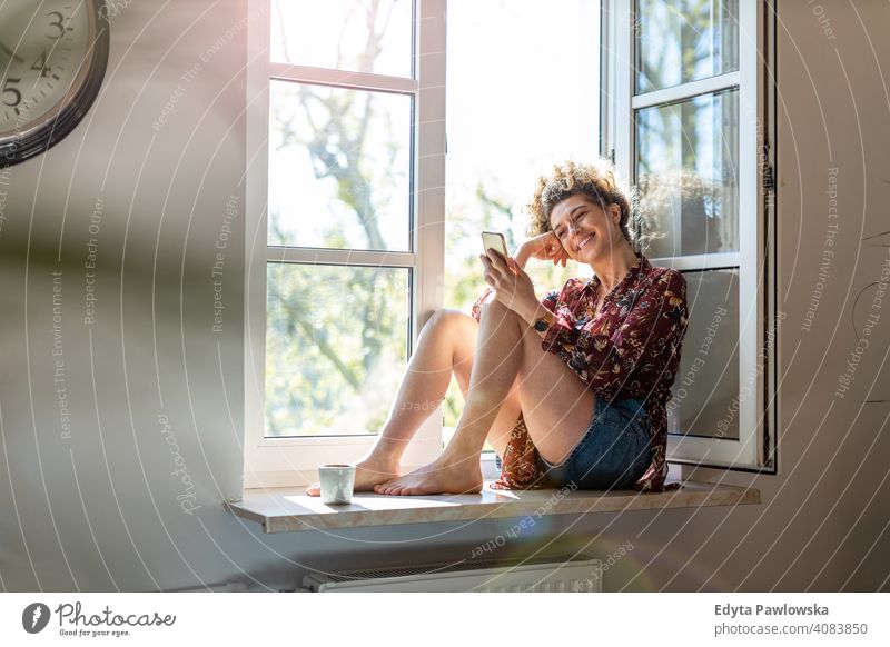 Young woman sitting on window sill and using smartphone happy smiling cheerful apartment leisure bedroom house home alone people caucasian adult indoors person