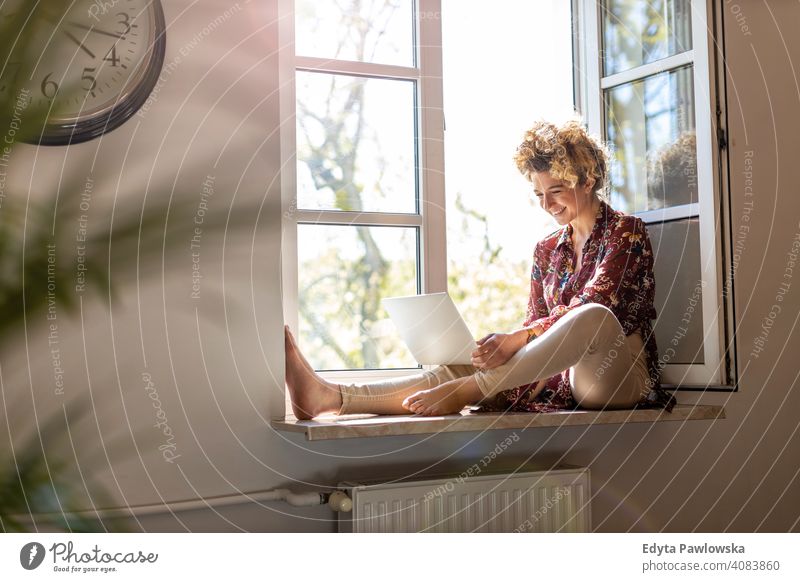 Young woman sitting on window sill and using laptop happy smiling cheerful apartment leisure bedroom house home alone people caucasian adult indoors person