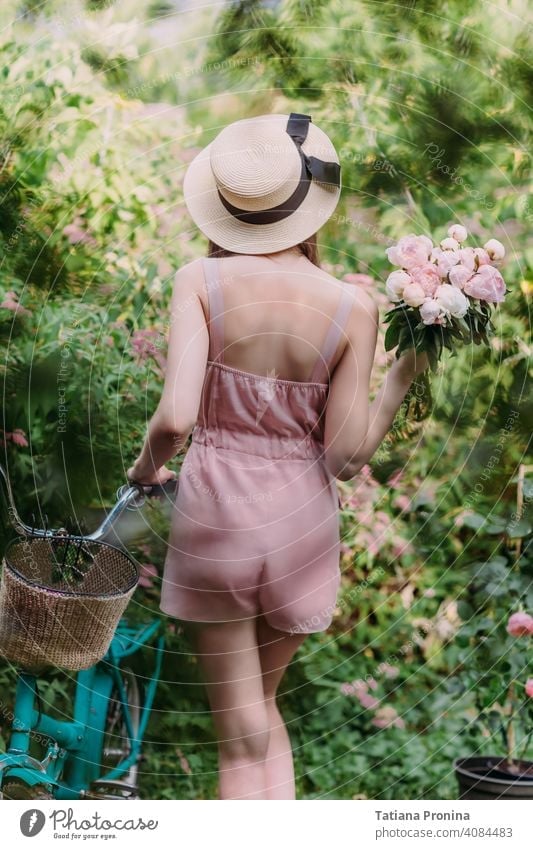 Stylish girl in a straw hat and a pale pink powdery jumpsuit walks with a mint-colored Bicycle in the garden of a country house. She holds a bouquet of fresh pink peonies in her hands. Stands with his back to the camera