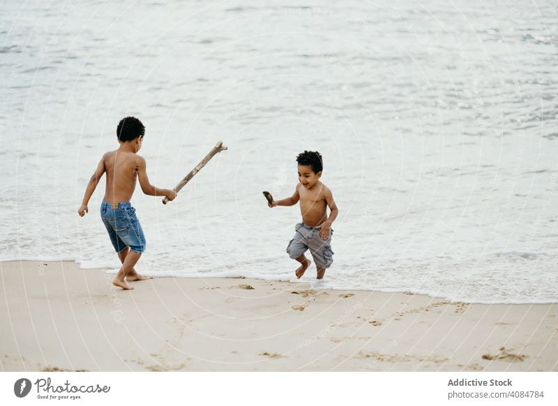 Black boys playing on beach brothers sea sticks fun together summer water shirtless barefoot kids children siblings game happy weekend lifestyle leisure shore