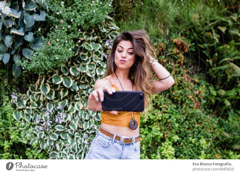 young blonde beautiful woman at the street taking a picture with mobile phone and smiling. Lifestyle outdoors. Summertime, Green background smart urban happy